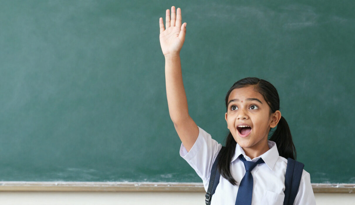 Young Indian girl in school uniform raising hand eagerly asking question, bright curious expression,