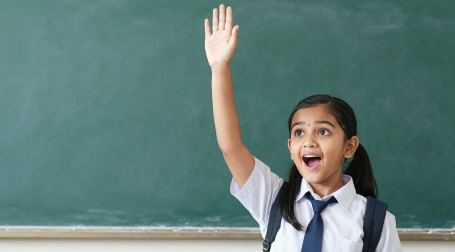 Young Indian girl in school uniform raising hand eagerly asking question, bright curious expression,